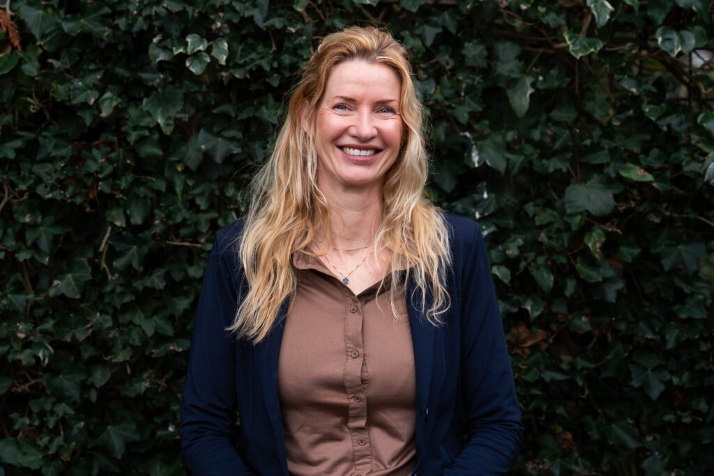 Vrouw met lange blonde haar die glimlacht, buiten voor een groene achtergrond.